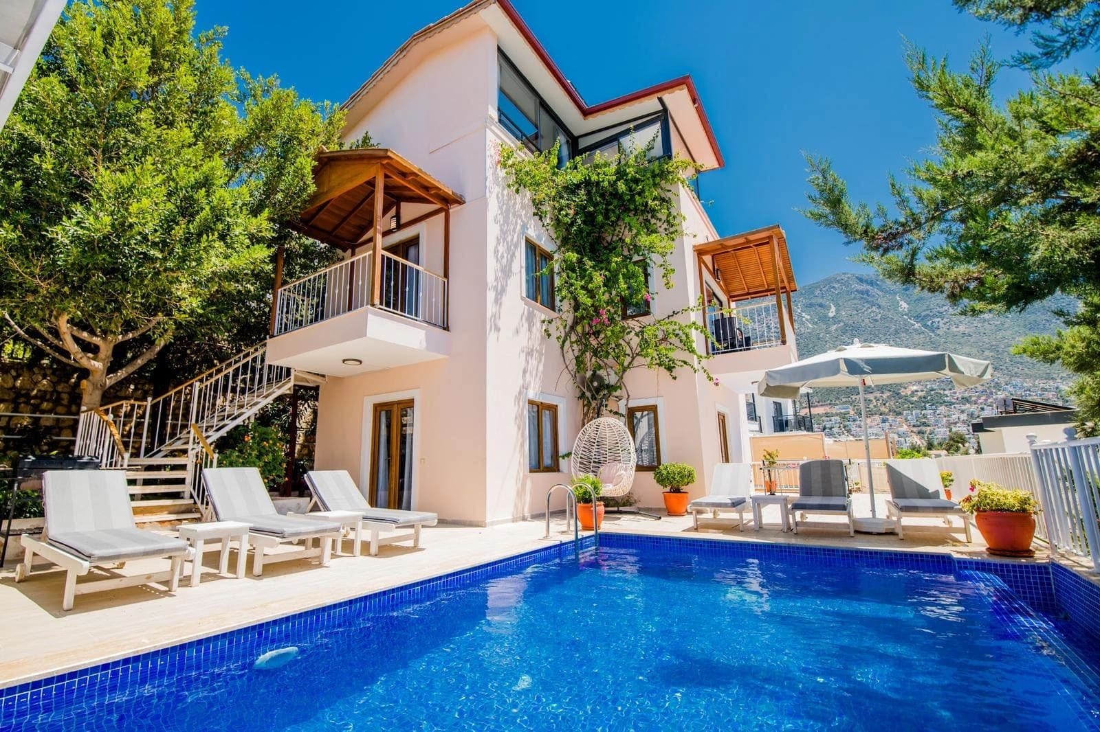 Açık pembe dış cepheli villa, önünde mavi havuz, şezlonglar ve asma şemsiye bulunan güneşlenme alanı.