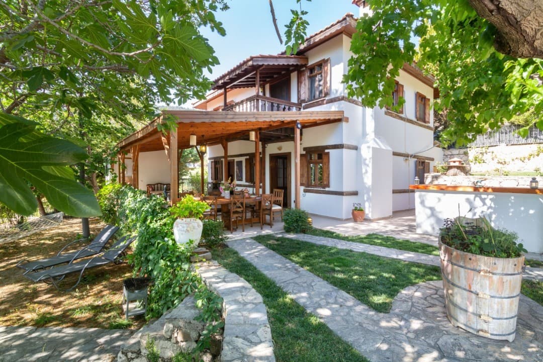 Ahşap detaylı iki katlı beyaz villanın önünde veranda, yemek masası ve çim bahçe yer alıyor.