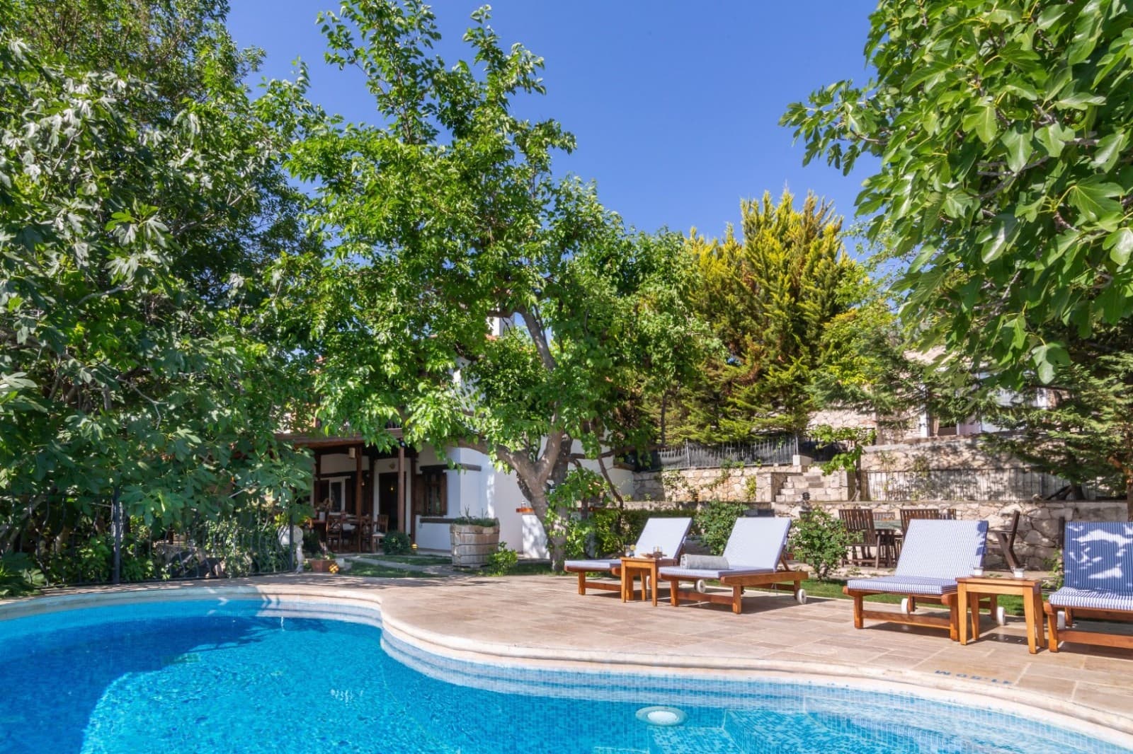 Ağaçların gölgelediği havuz kenarında şezlonglar ve arka planda beyaz villa.