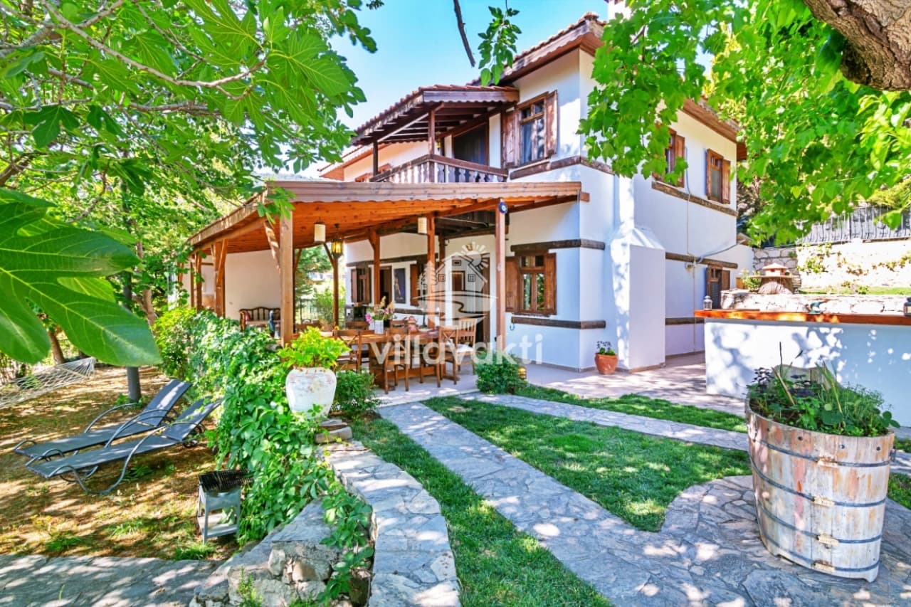 Taş duvarlı, ahşap detaylı villa bahçesinde gölgelikli yemek masası, şezlonglar ve yeşil bitkiler.