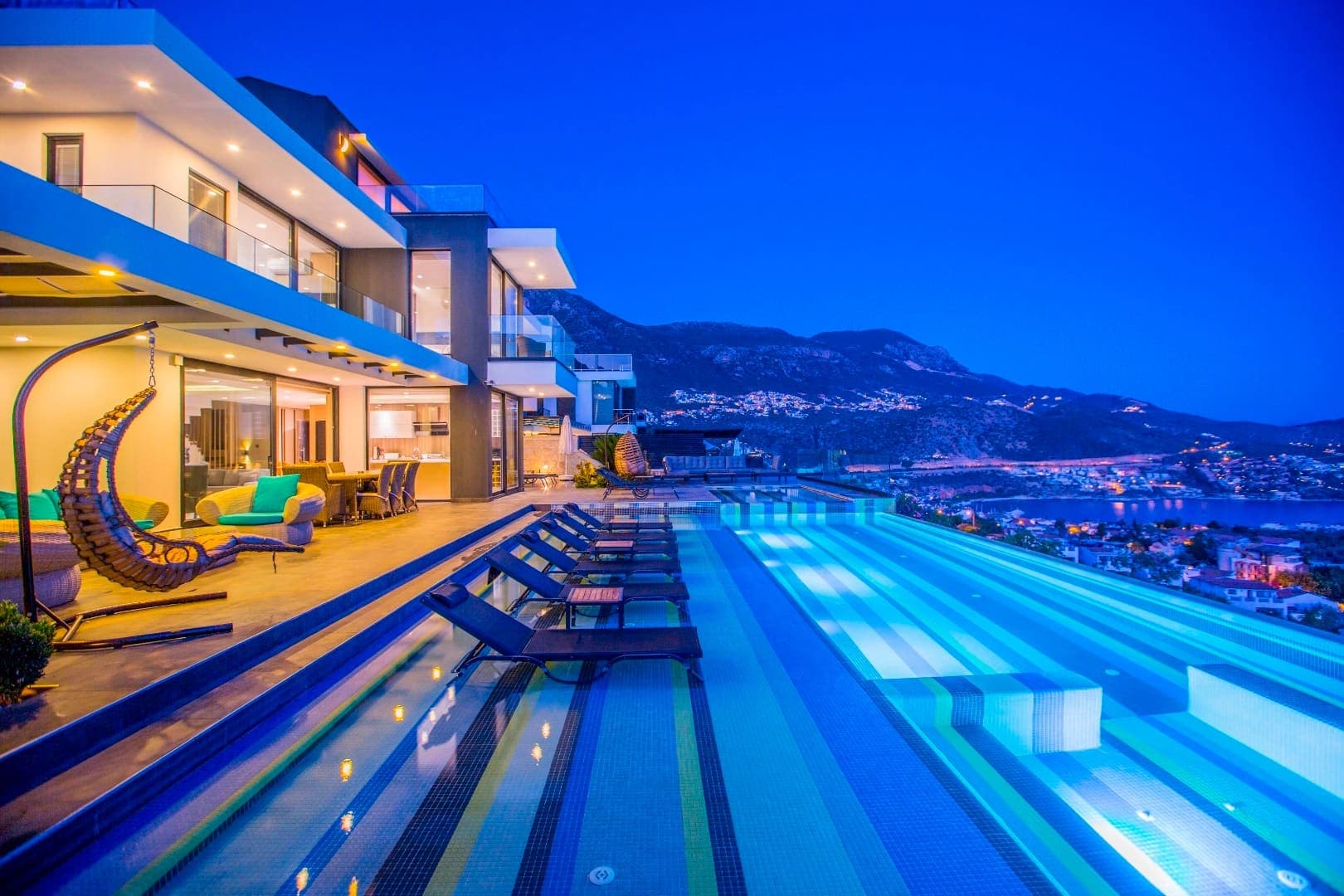 Gece aydınlatmalı sonsuzluk havuzu, şezlonglar ve modern villanın dış cephesi.