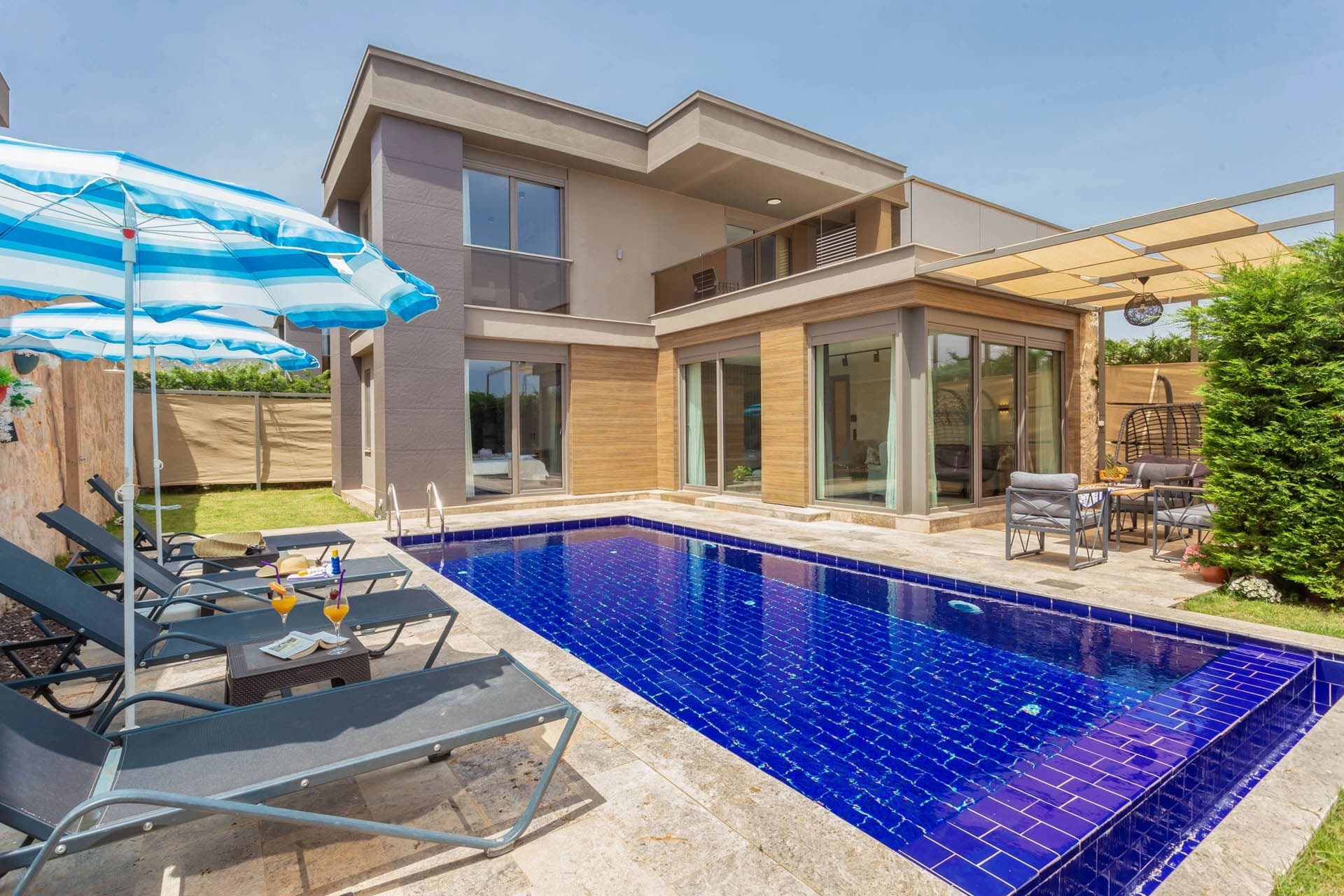 Mavi şemsiyeli şezlongların bulunduğu havuz kenarı ve modern iki katlı villa dış cephesi.