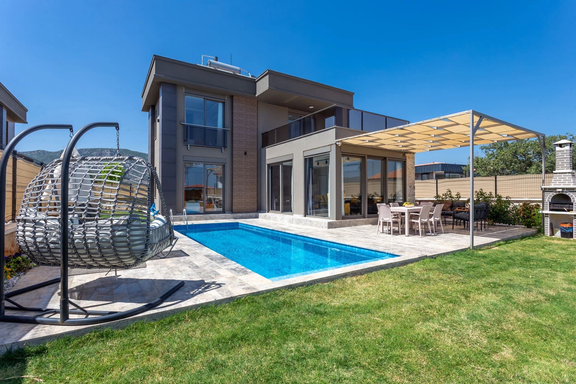Bahçesinde salıncak, havuz, yemek masası ve taş barbekü bulunan modern iki katlı villa.