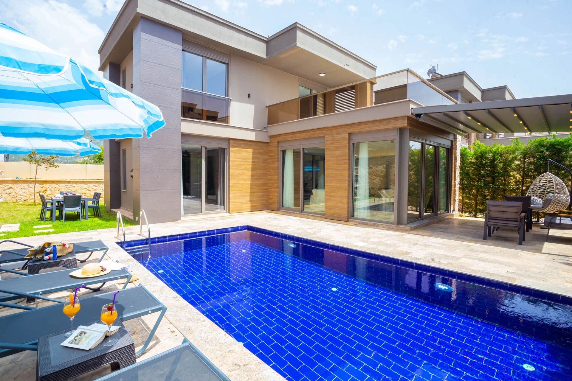 Mavi havuz başında şezlonglar, şemsiye ve modern dış cepheye sahip villa.