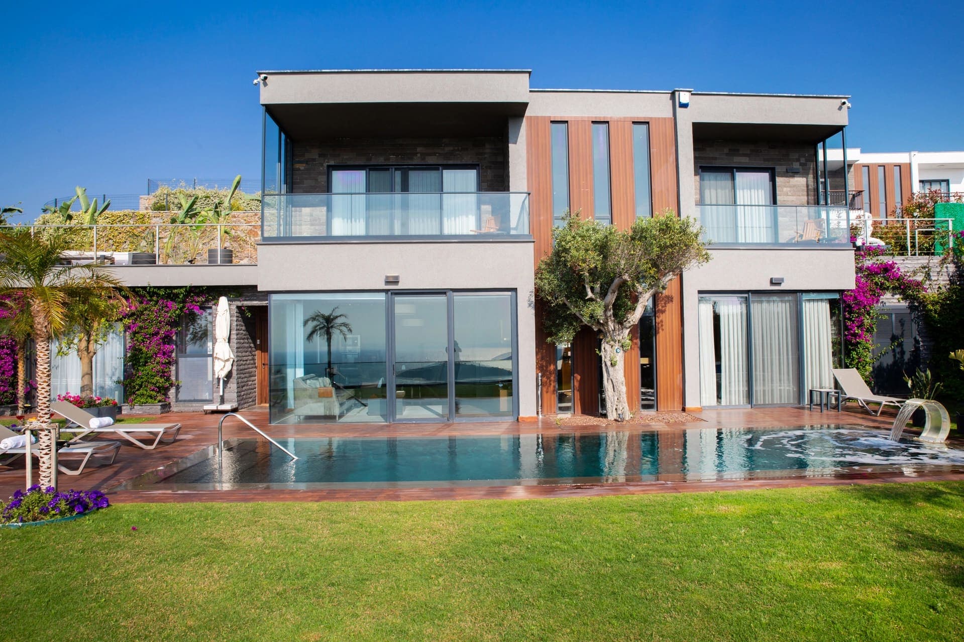 Önünde havuz, yeşil bahçe ve ağaç bulunan modern, iki katlı taş ve ahşap detaylı villa.