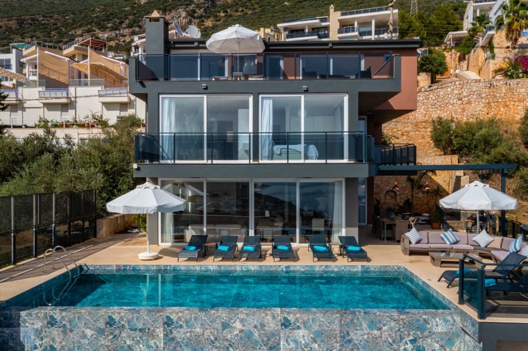 Önünde yüzme havuzu, şezlonglar ve oturma grubu bulunan, balkonlu, modern gri dış cepheli villa.