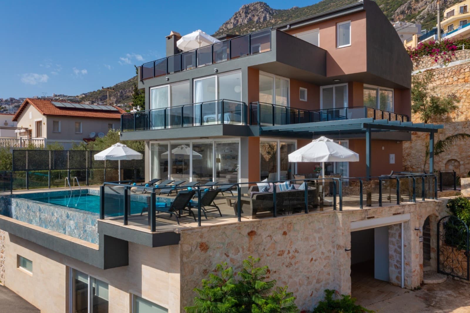 Dağ manzaralı, çok katlı ve kahverengi tonlarında modern villa, önünde havuz ve şezlonglar var.