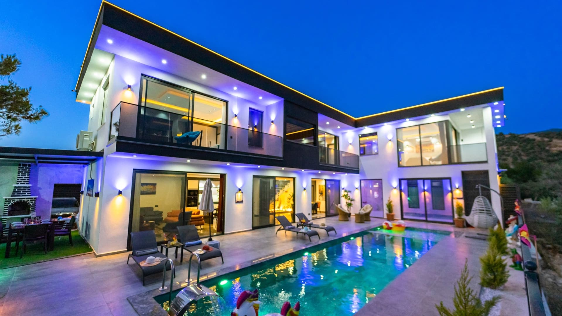 Akşam vakti aydınlatılmış, havuz başında şezlonglar ve oturma grubu bulunan modern villa dış cephesi.