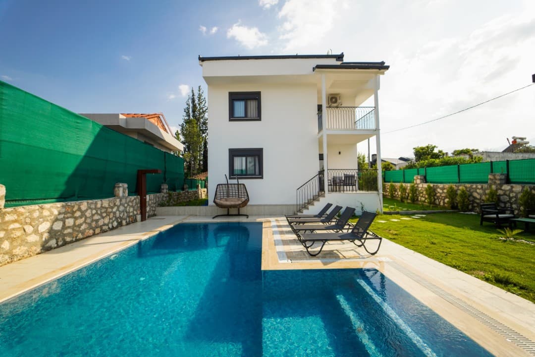 Önünde yüzme havuzu, şezlonglar ve asma koltuk bulunan iki katlı beyaz dış cepheli villa.