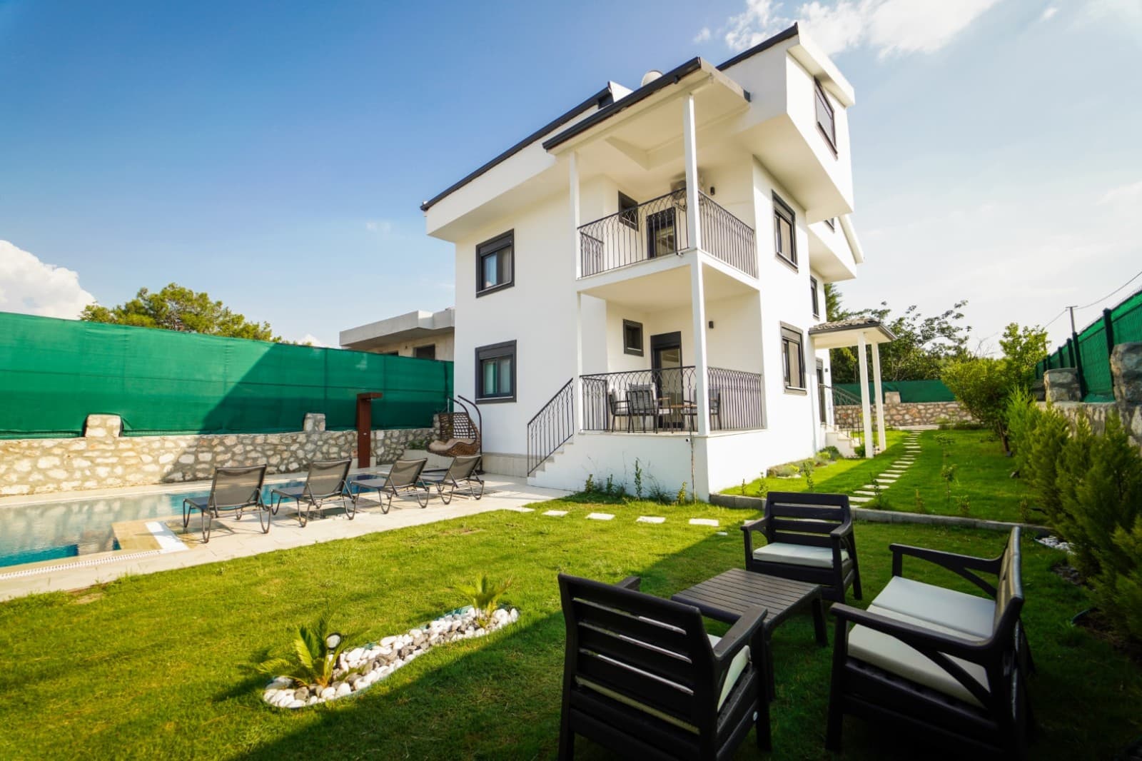 İki katlı beyaz villa, önünde çim alan, havuz ve bahçe oturma grubu bulunuyor.