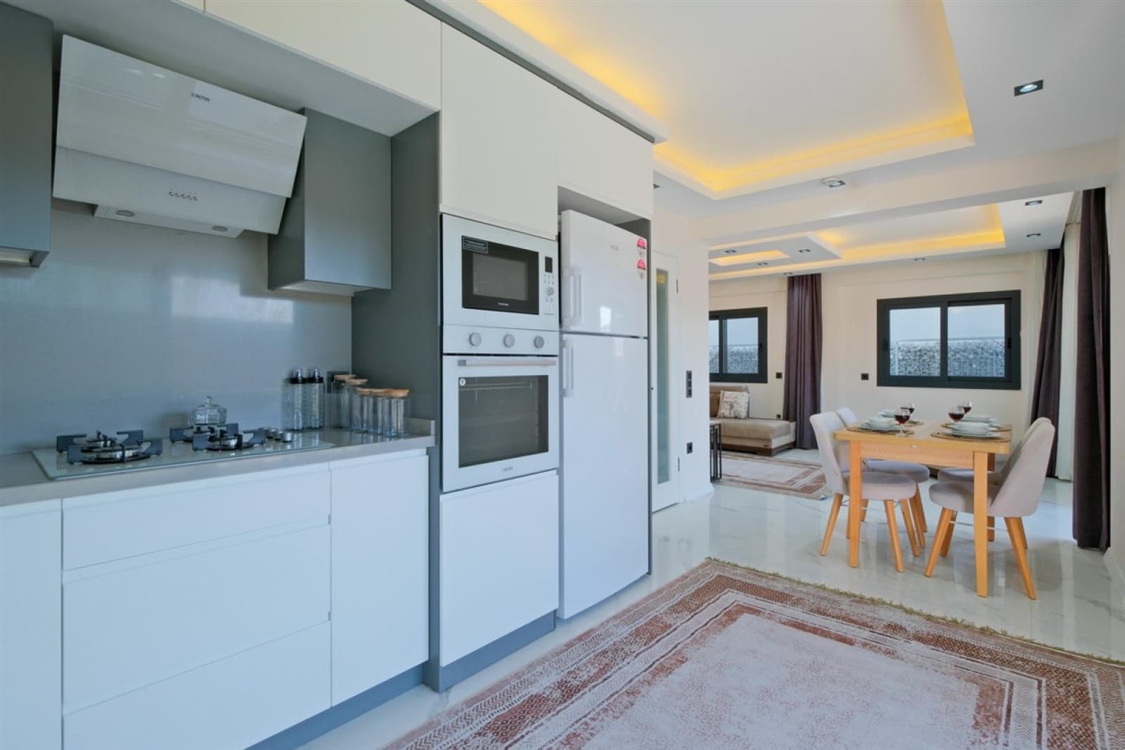Modern beyaz mutfak dolapları, ankastre fırın, buzdolabı ve yemek masası.