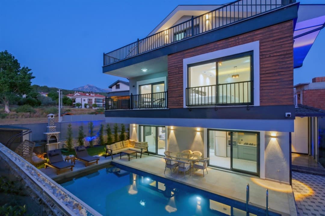 Akşam karanlığında havuzu, oturma grubu ve ışıklandırılmış dış cephesi görünen çok katlı villa.