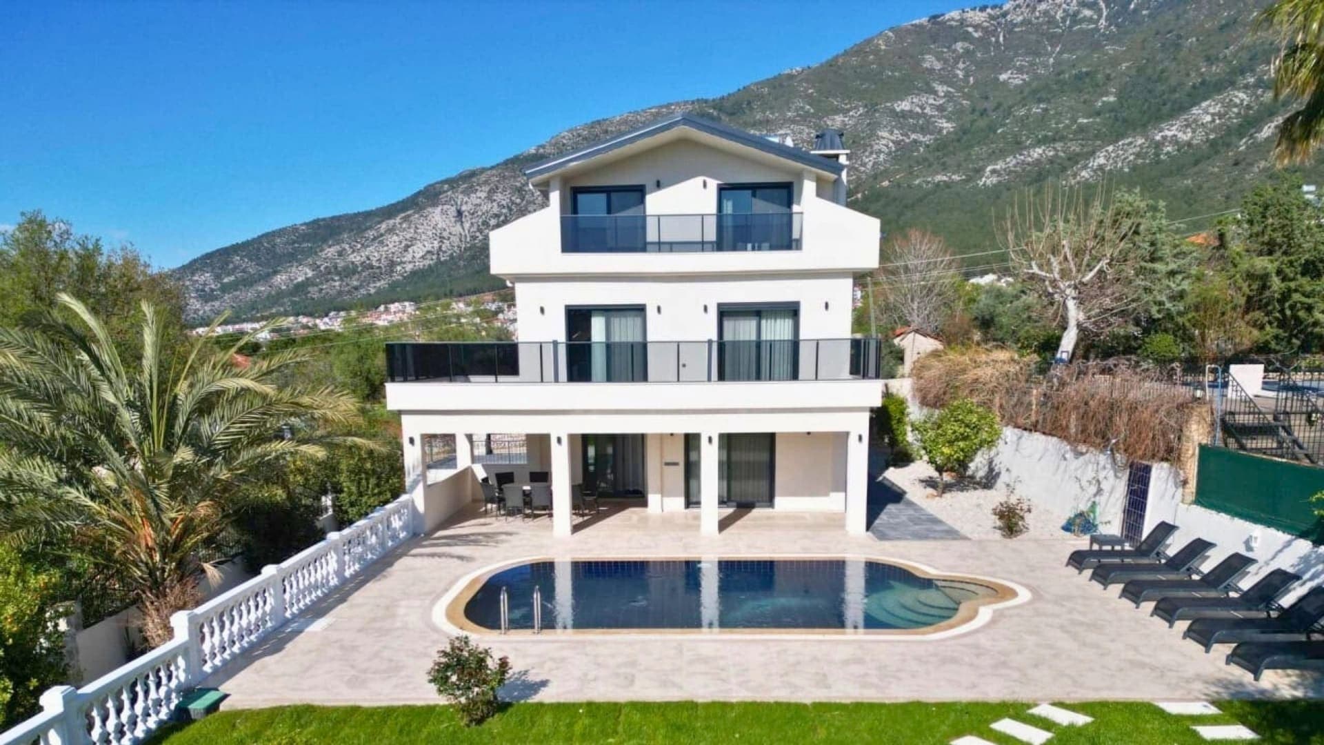 Dağ manzarası önünde üç katlı beyaz villa, açık yüzme havuzu, çim bahçe ve şezlonglar.