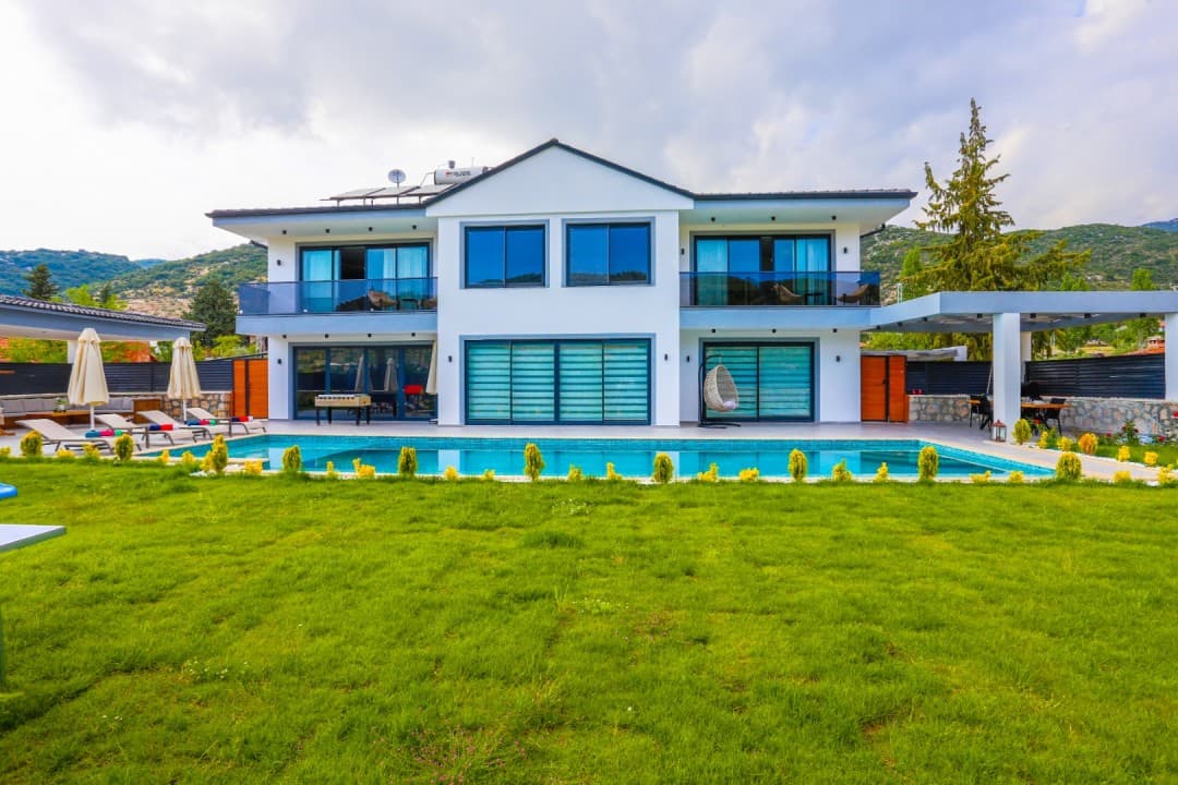 Önünde havuz ve yeşil bahçe bulunan, iki katlı, beyaz dış cepheli ve balkonlu müstakil villa.