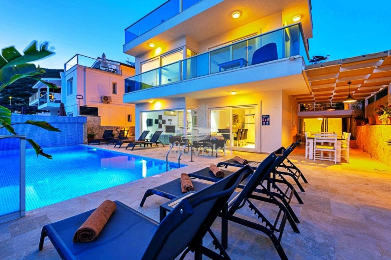 Akşam ışıklandırmalı modern villa, havuz kenarında şezlonglar ve yemek alanı mevcut.