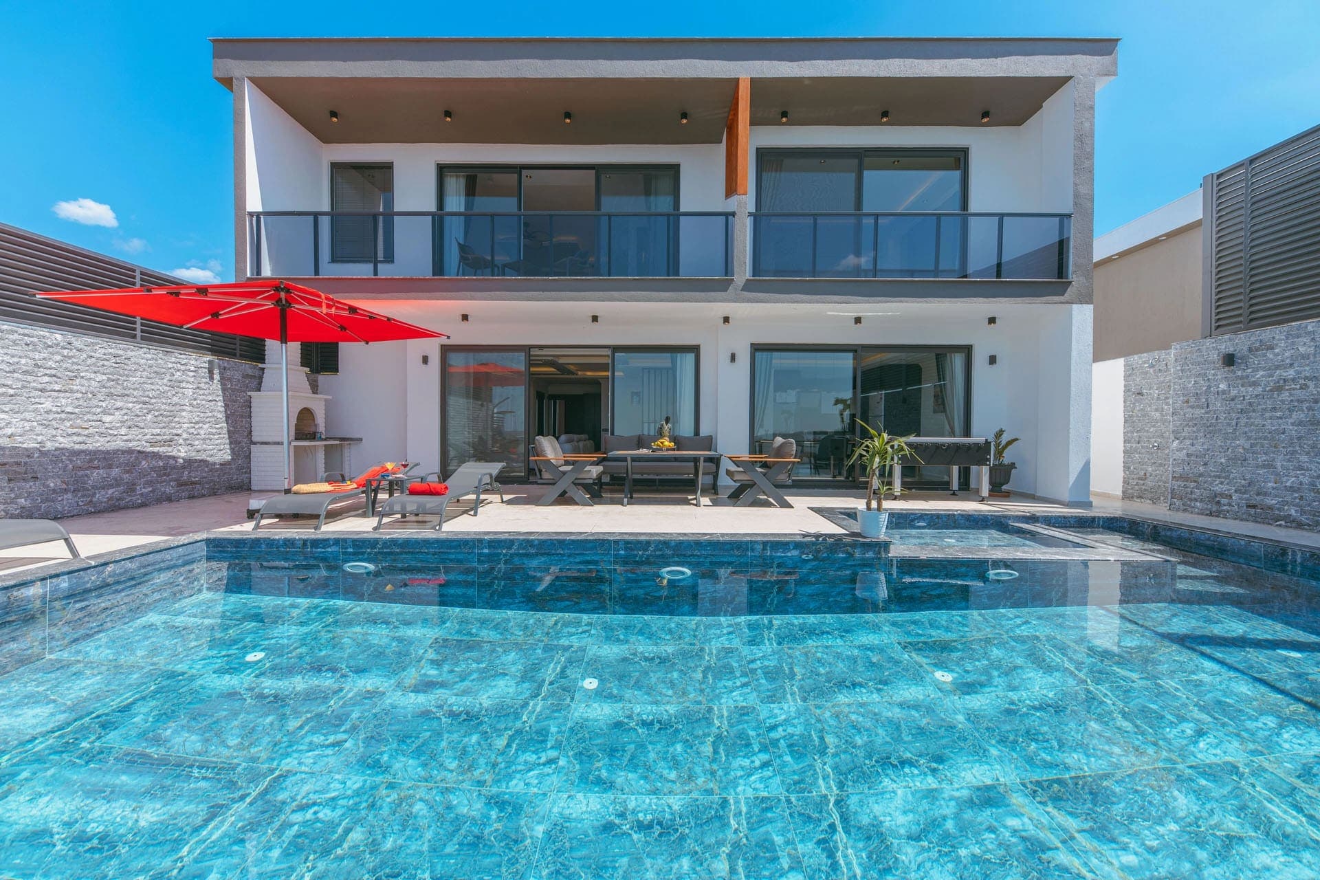 İki katlı modern villa, önünde mavi mozaik kaplı havuz, kırmızı şemsiye ve oturma grubu bulunuyor.