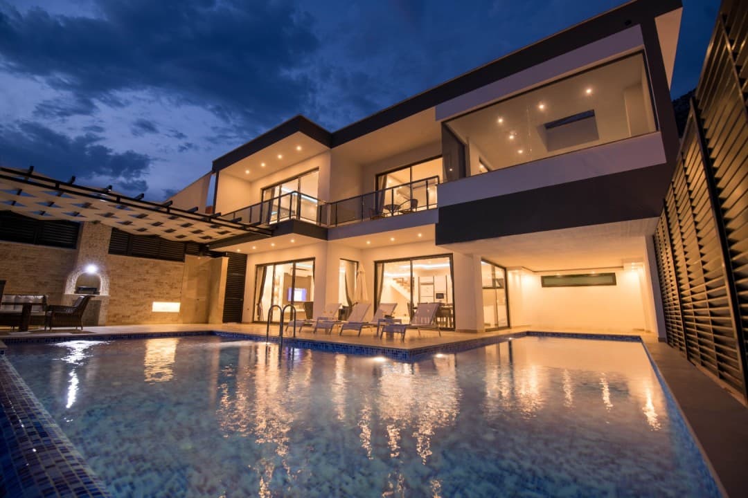Akşam vakti aydınlatılmış modern villanın havuzlu dış cephesi.