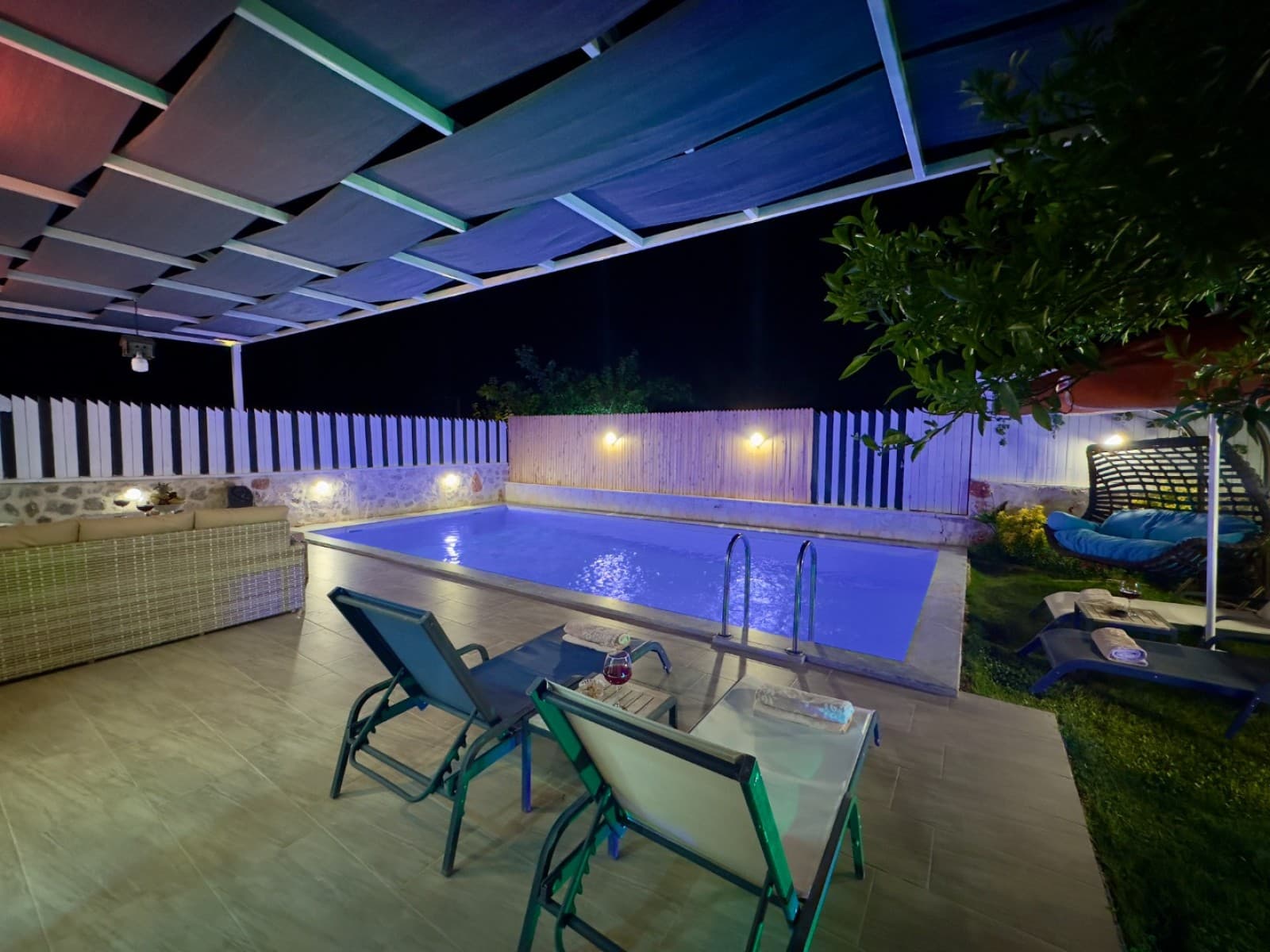 Gece aydınlatmalı havuz kenarında şezlonglar ve tenteli oturma alanı.