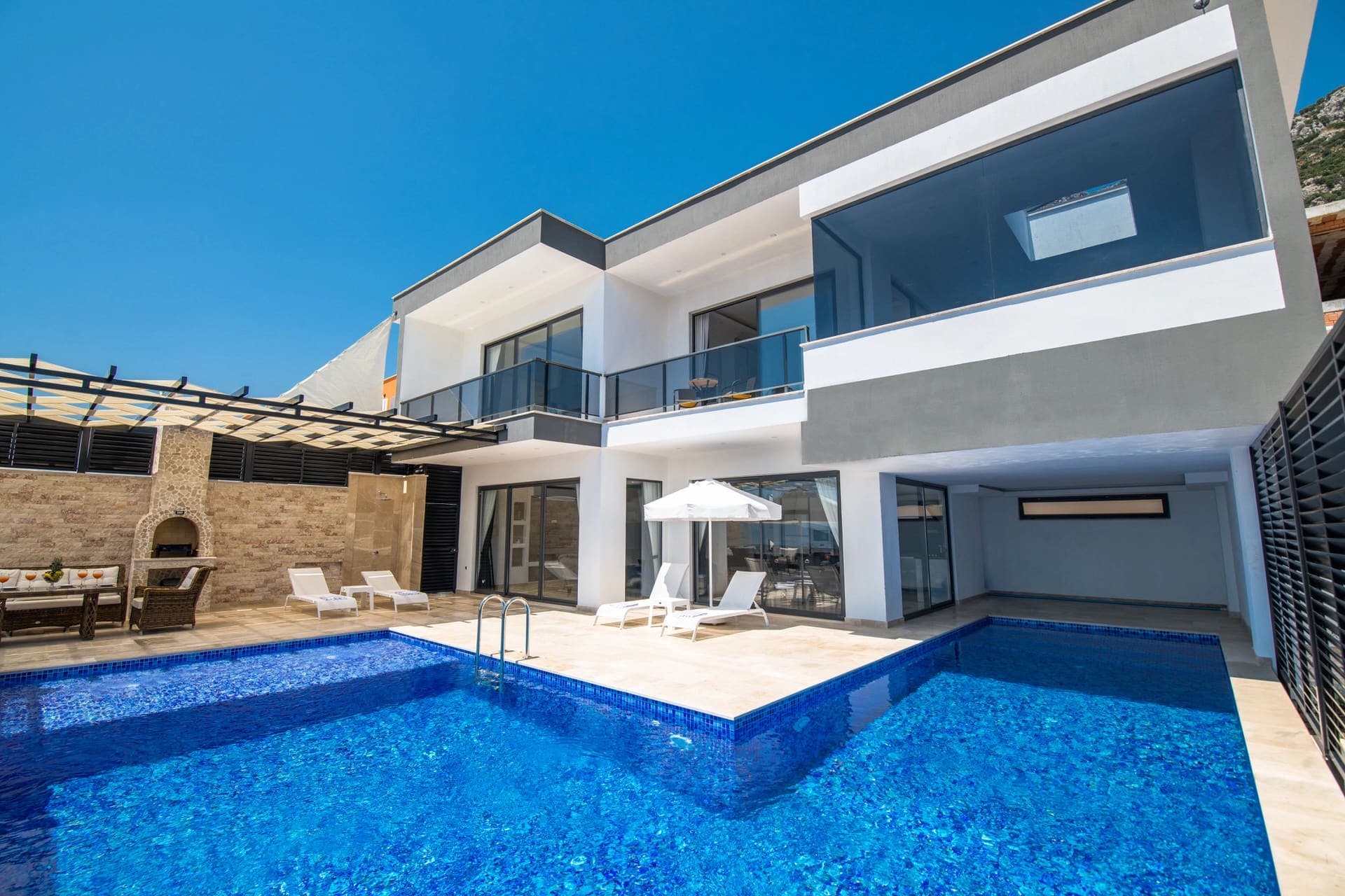 İki katlı, beyaz ve gri tonlarda modern villa, önünde özel yüzme havuzu ve şezlonglar bulunuyor.