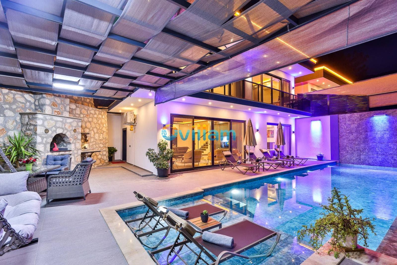 Akşam aydınlatmalı, havuz başında şezlonglar ve taş duvarlı barbekü alanı bulunan modern villa.