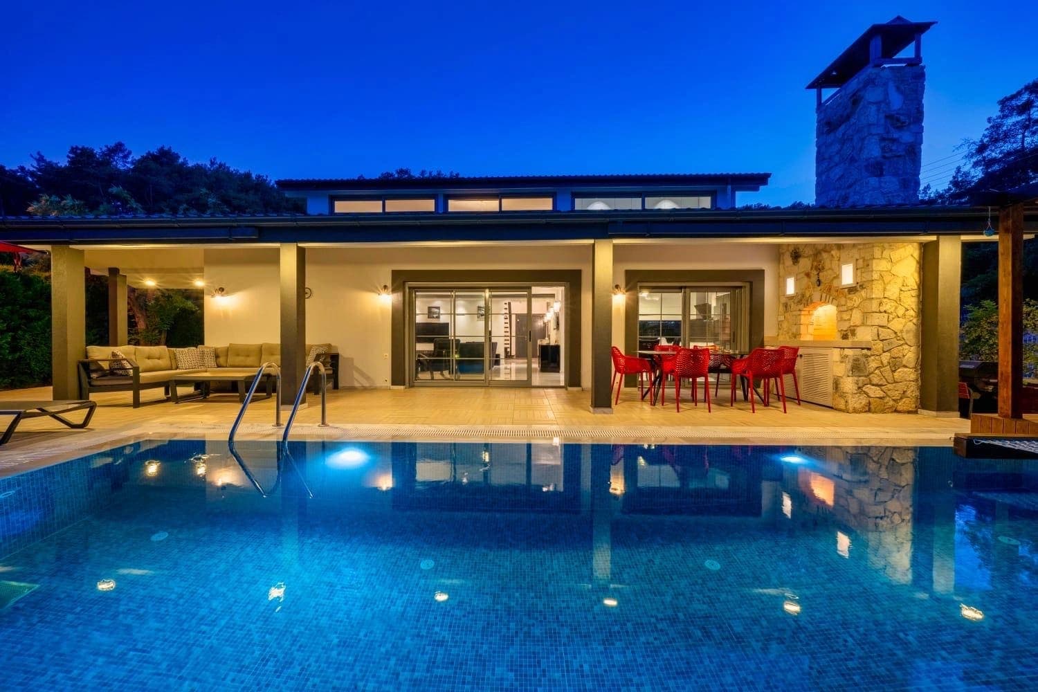 Gece aydınlatmalı havuz, terasta yemek masası ve oturma grubu bulunan villa dış cephesi.