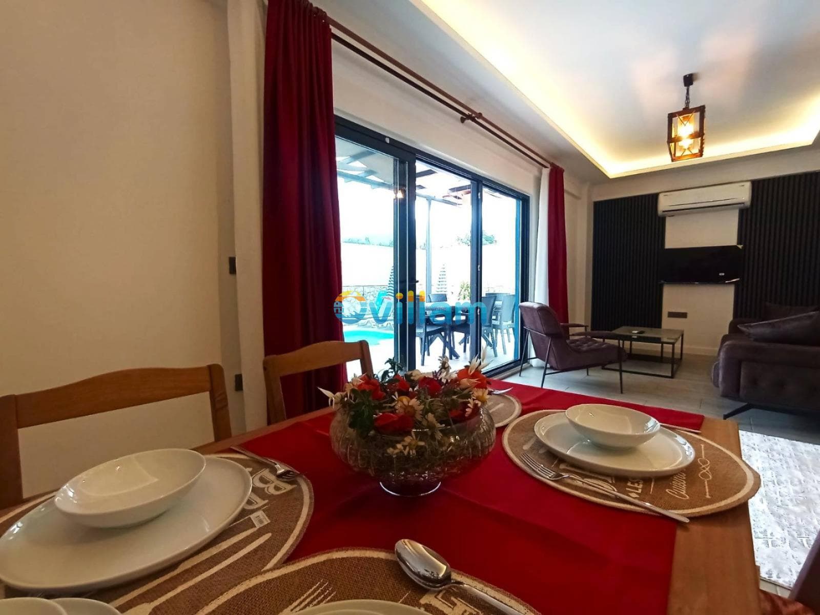 Kırmızı örtülü yemek masası, servis tabakları ve arka planda havuz manzaralı oturma alanı.