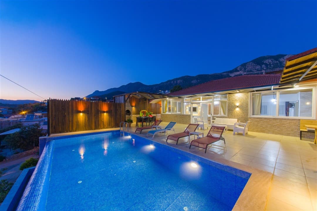 Akşam karanlığında havuz kenarındaki şezlonglar ve arka planda dağ manzarası olan villa dış cephesi.