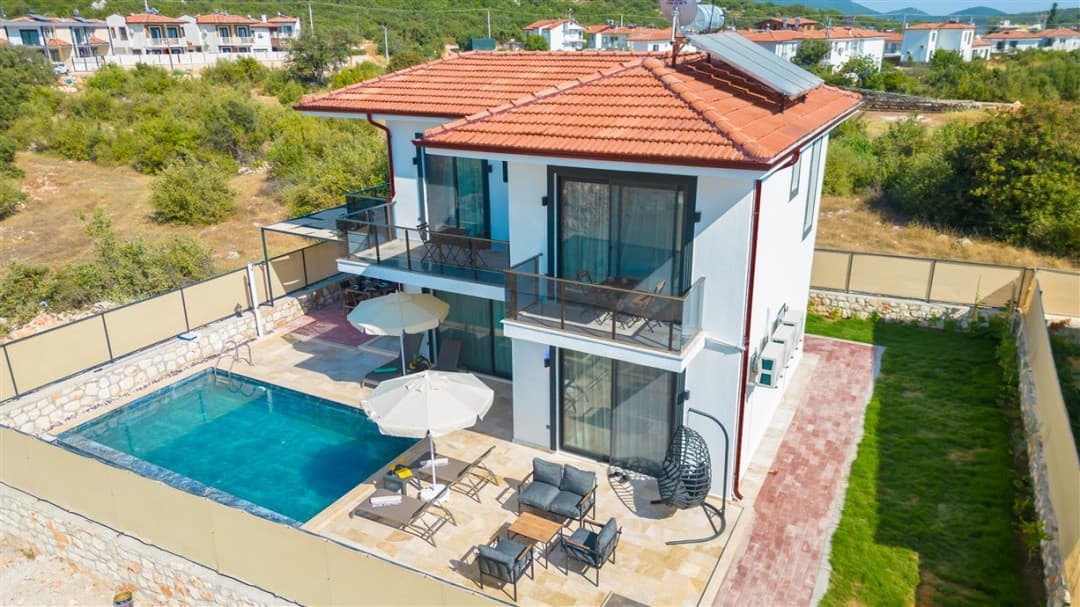 Kiremit çatılı, iki katlı, balkonlu beyaz villanın önünde özel havuz, şezlonglar ve oturma grubu bulunuyor.