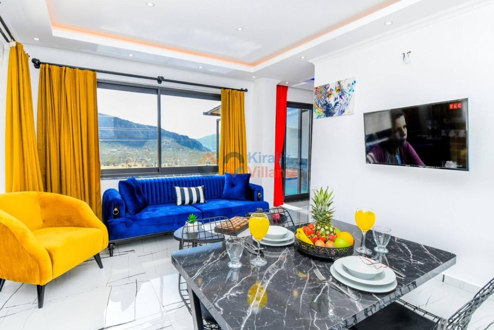 Mavi koltuk, sarı berjer, televizyon ve mermer desenli yemek masası bulunan salon.