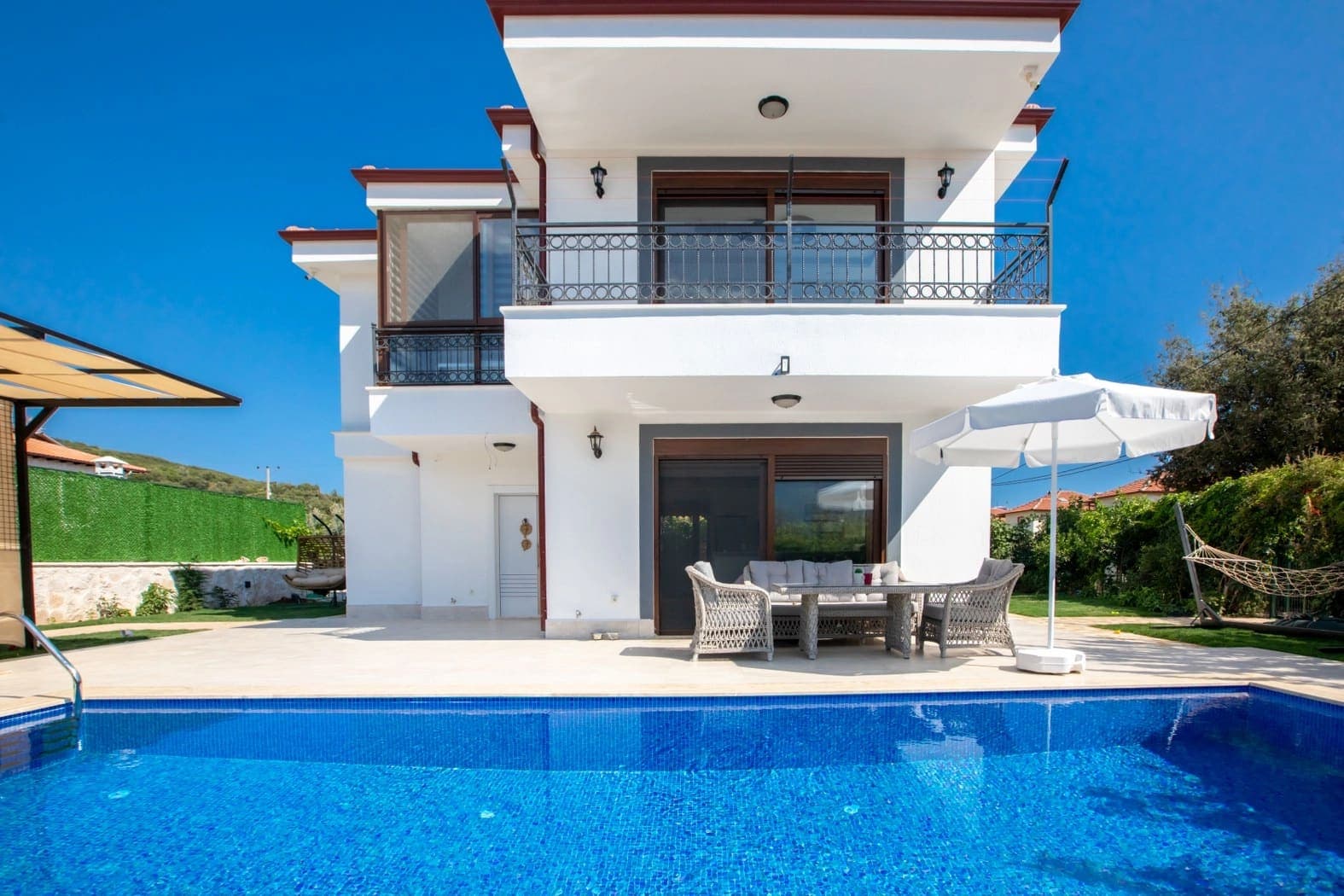 Önünde özel yüzme havuzu ve şemsiyeli oturma grubu bulunan iki katlı beyaz villa.