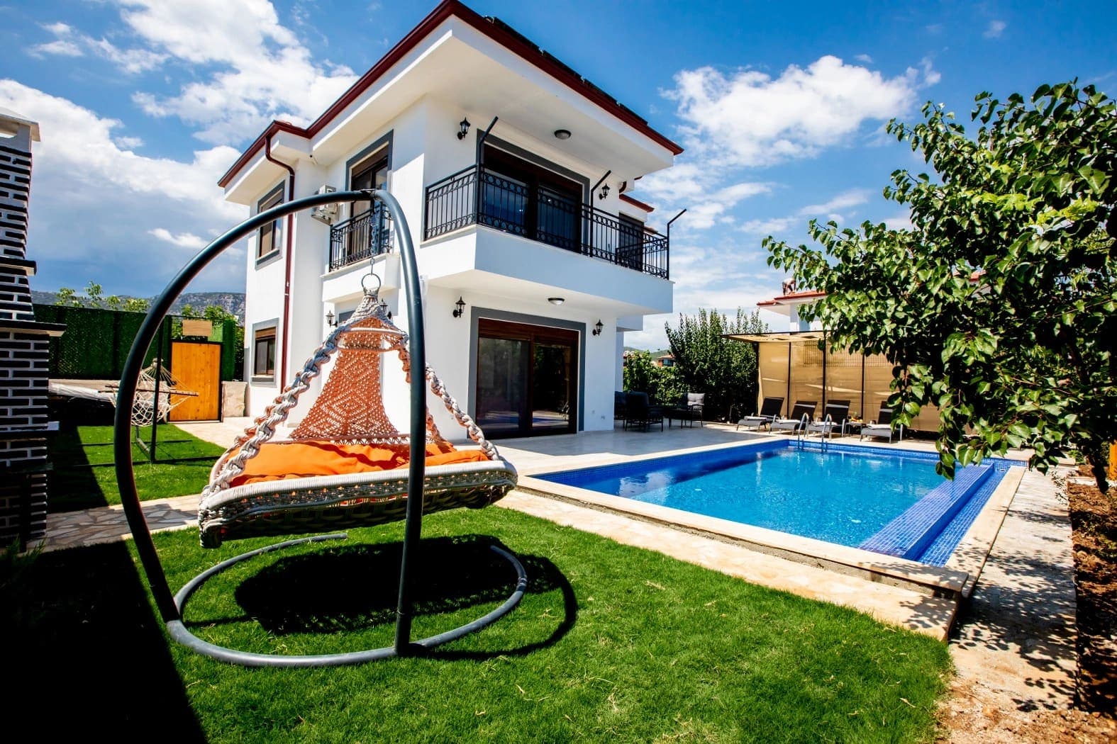 Çim bahçesinde makrome salıncak, yüzme havuzu ve şezlonglar olan iki katlı villa.
