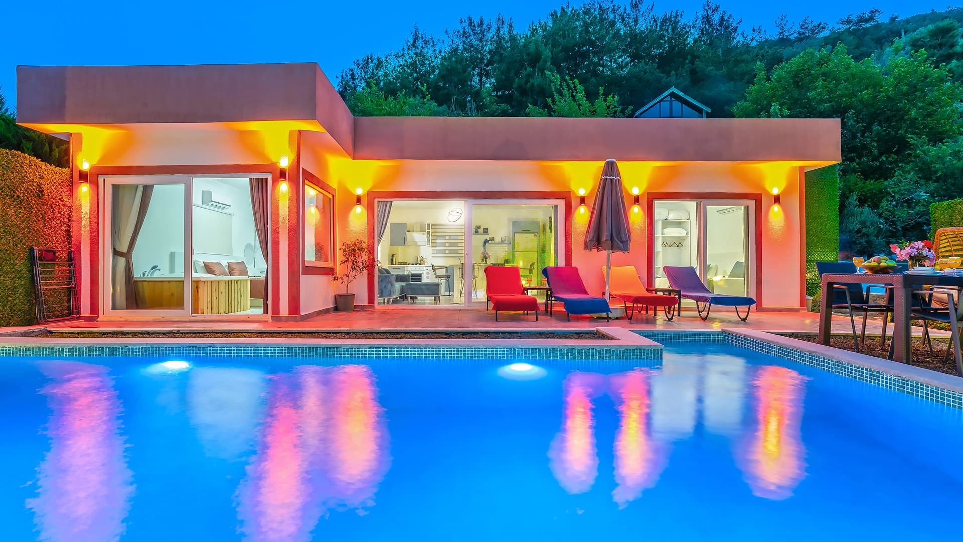 Gece aydınlatmalı yüzme havuzu önünde şezlonglar ve ışıklandırılmış villa dış cephesi.