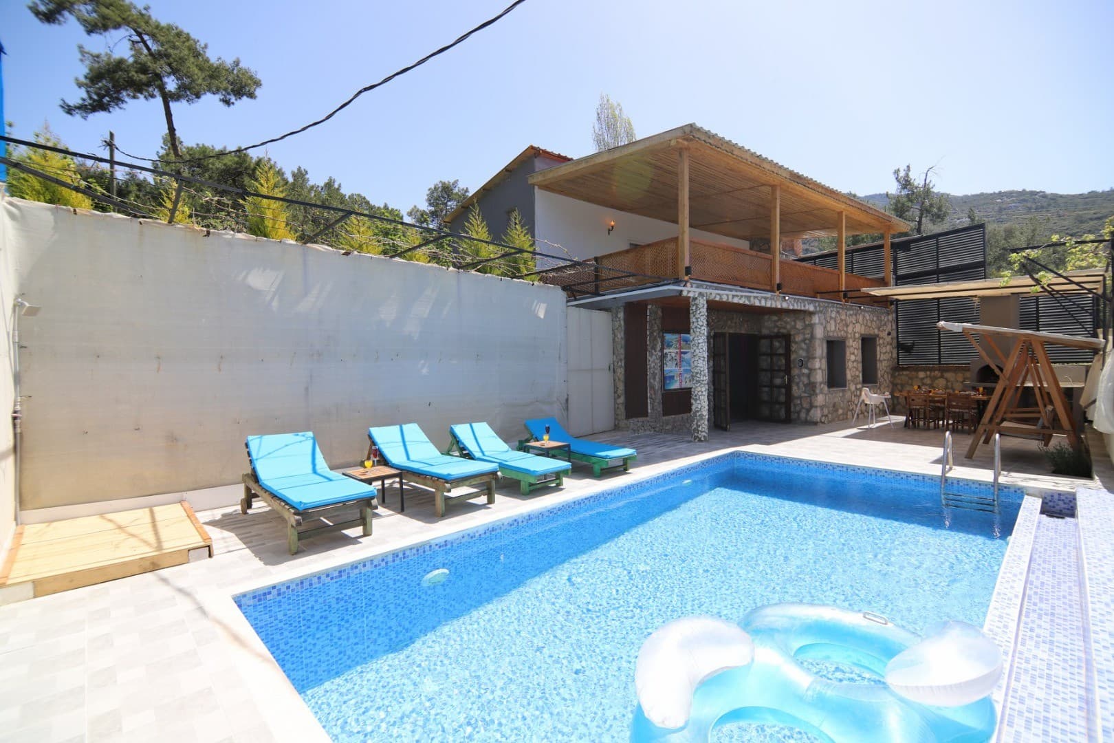 Mavi havuzlu, şezlonglu, ahşap sundurmalı ve taş duvarlı iki katlı villa bahçesi.