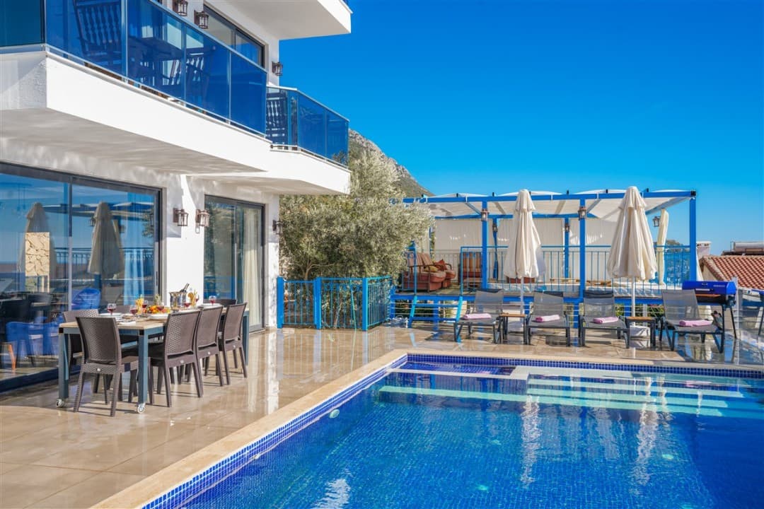 Gündüz vakti havuz kenarında yemek masası ve şezlonglar bulunan beyaz villa.