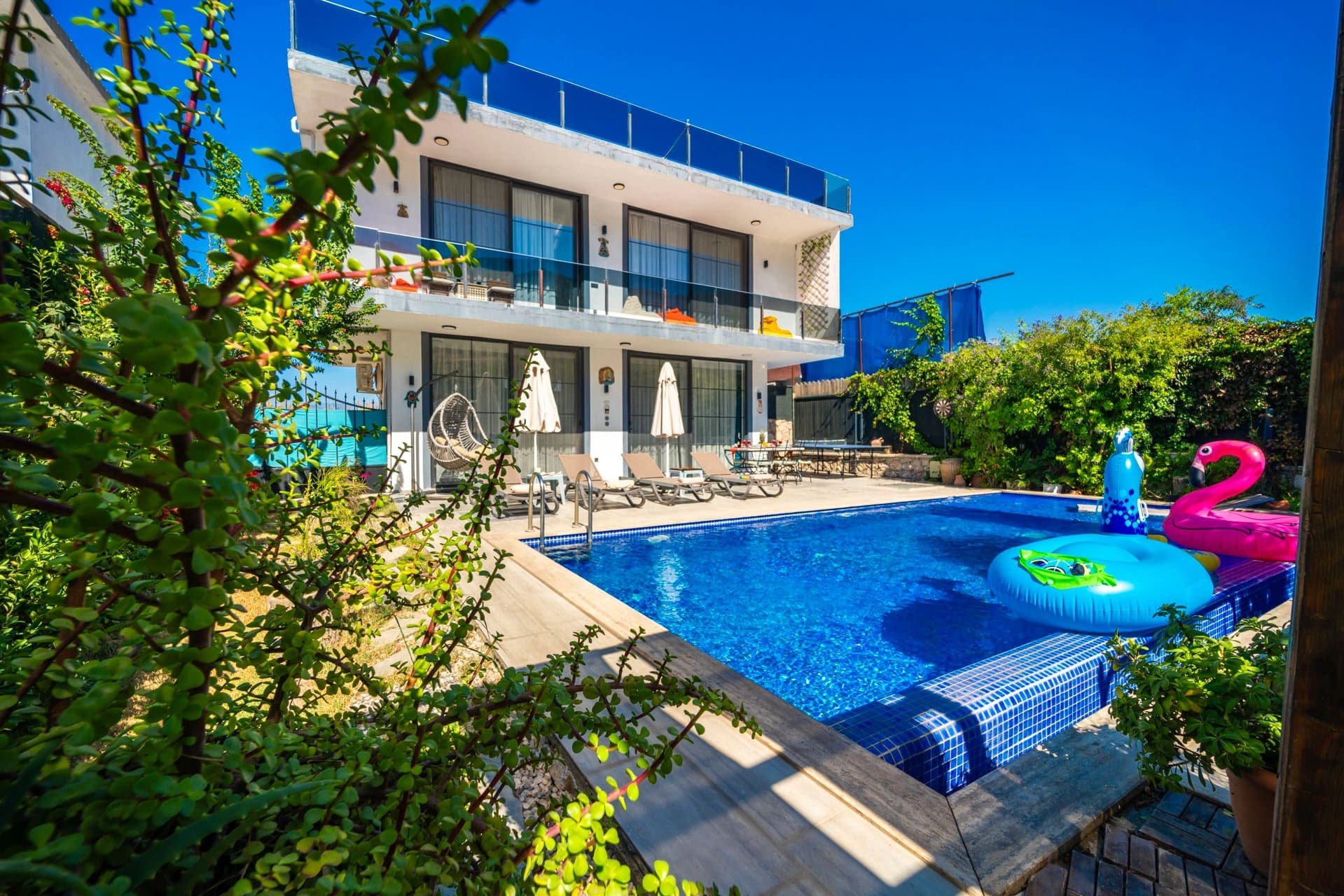 İki katlı modern villa, önünde havuz ve şişme deniz oyuncakları.