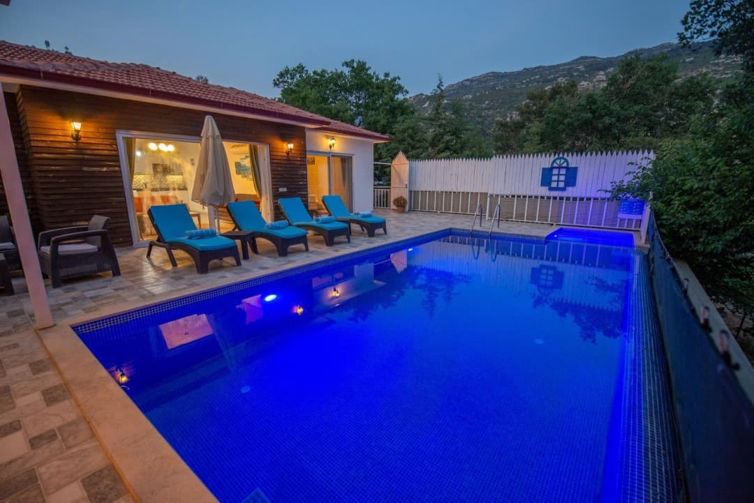 Aydınlatmalı mavi havuzun başında şezlonglar ve ahşap cepheli villa.