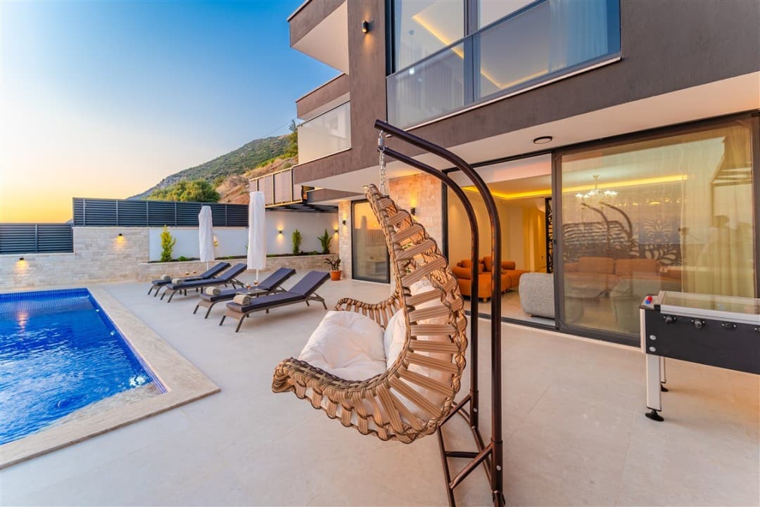 Havuz kenarında hasır salıncak, şezlonglar ve arka planda modern bir villa bulunan gün batımı manzarası.