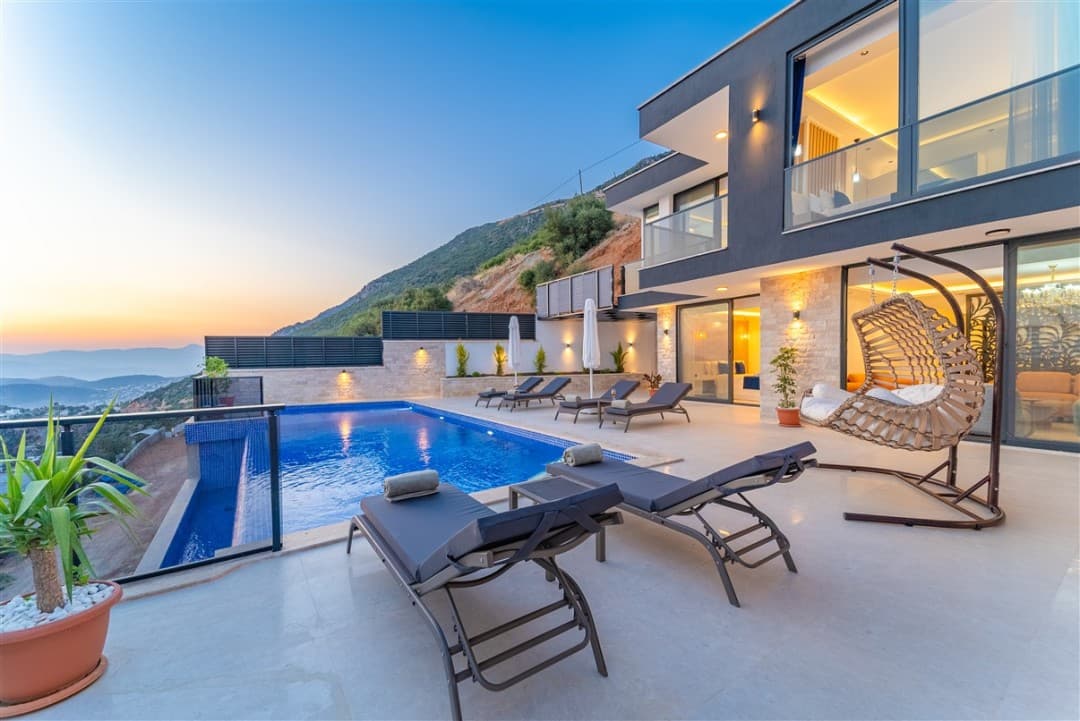 Gün batımında dağ manzaralı havuz başı ve şezlongları olan modern iki katlı villa.
