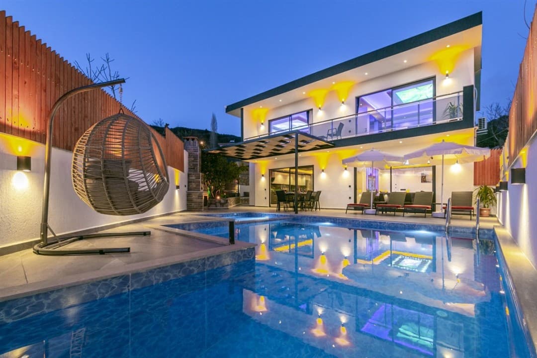 Akşamüstü aydınlatmalı havuz kenarında salıncak ve şezlonglar bulunan iki katlı villa.