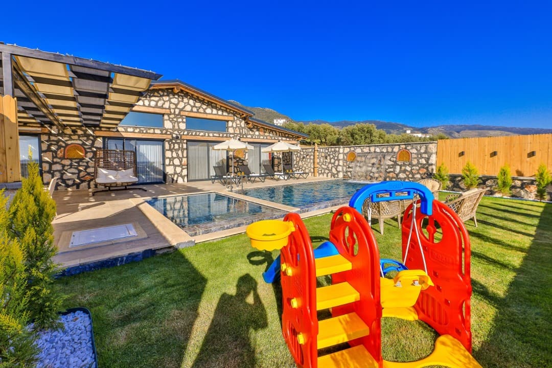Çim alanda renkli çocuk kaydırağı ve arkada havuzlu villa.
