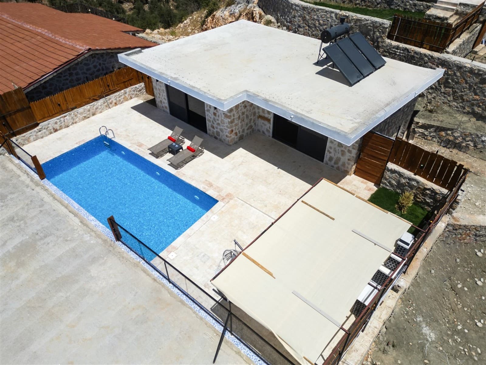 Havuz kenarında şezlonglar bulunan, düz çatılı ve güneş panelli müstakil taş villa.