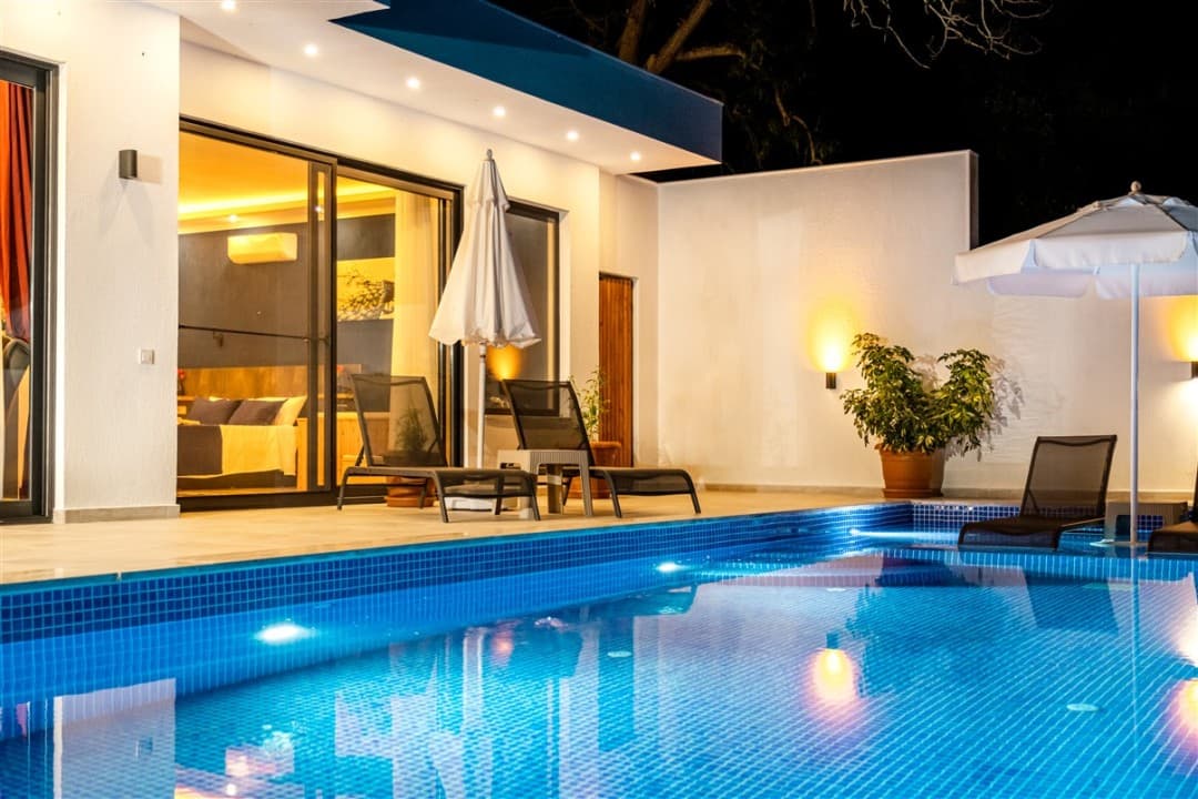 Geceleri havuz başı aydınlatmalı, şezlonglu ve şemsiyeli villa terası.