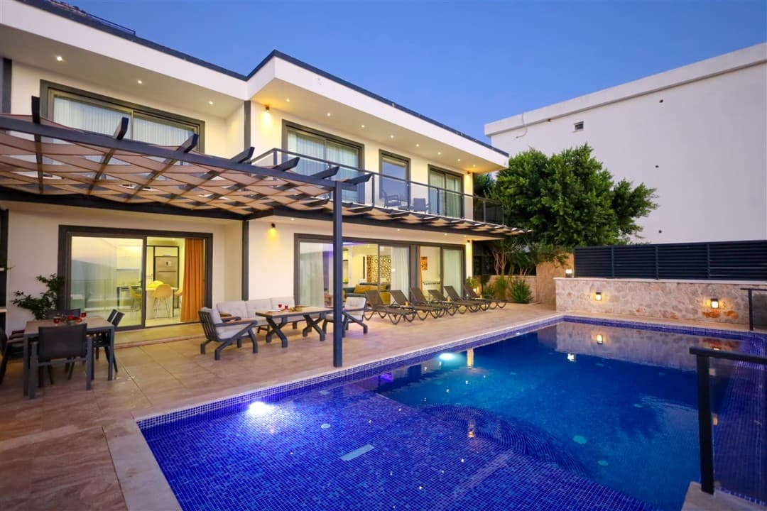 Akşam ışıklandırmalı, havuz kenarında oturma grubu ve şezlonglar bulunan iki katlı modern villa.
