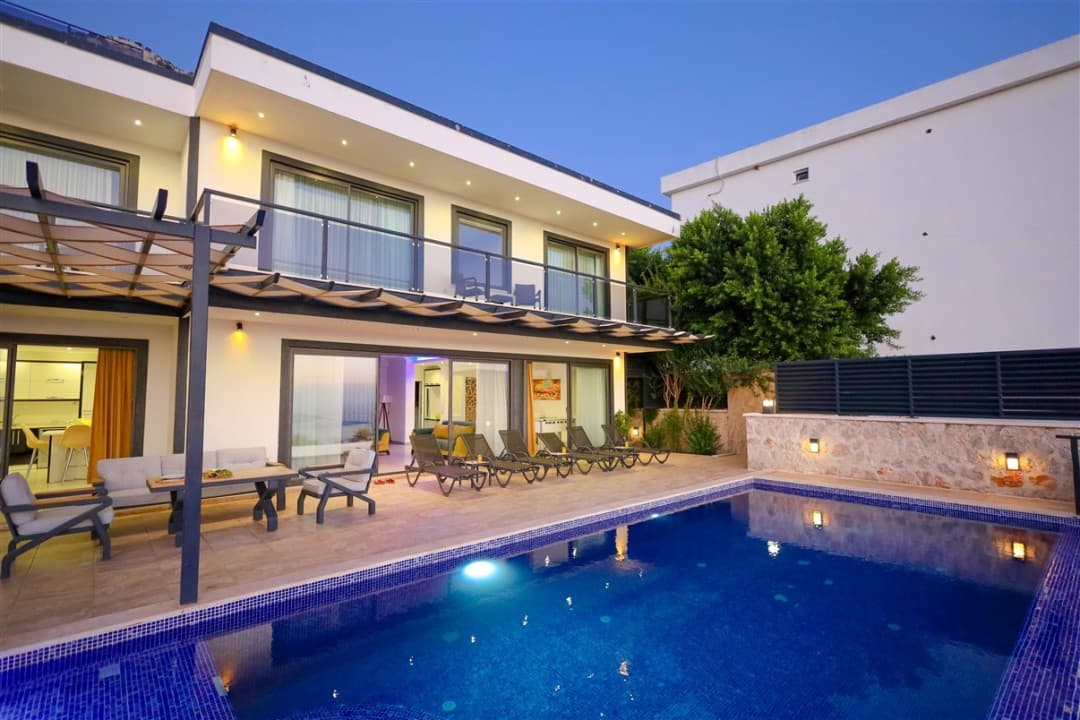 Akşamüstü aydınlatmalı havuz kenarında şezlonglar ve iki katlı modern villa.