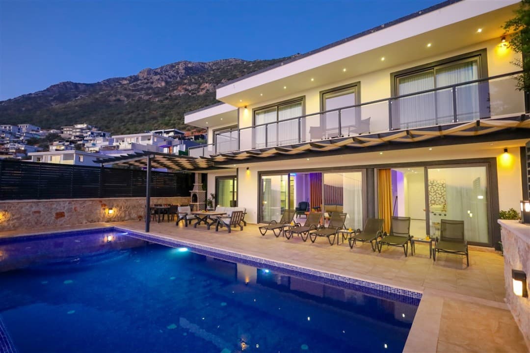Akşam vakti havuz başı şezlonglu ve dağ manzaralı lüks villa dış cephesi.
