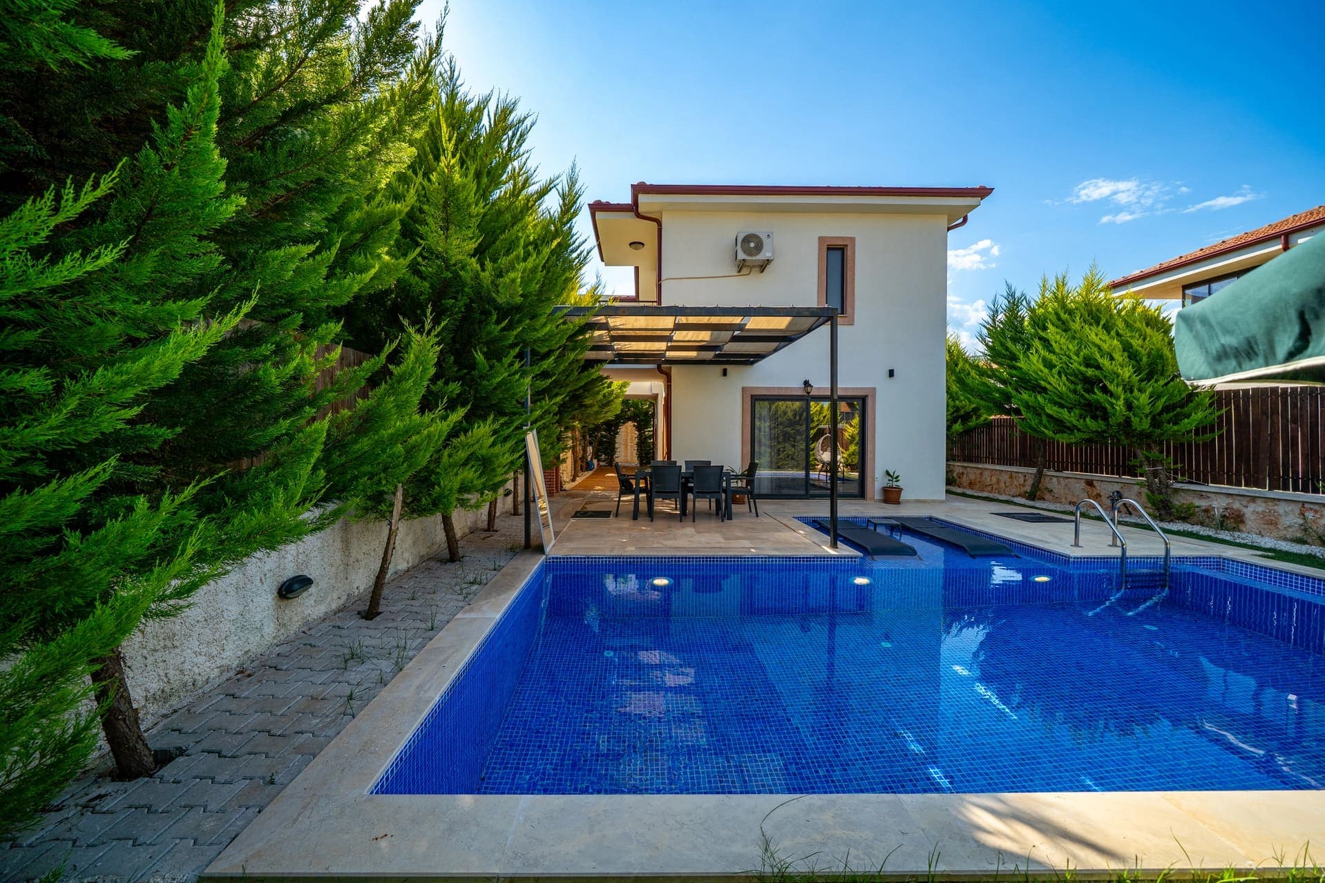 Özel yüzme havuzu, havuz kenarında şezlonglar ve ağaçlarla çevrili bahçesi olan iki katlı villa.