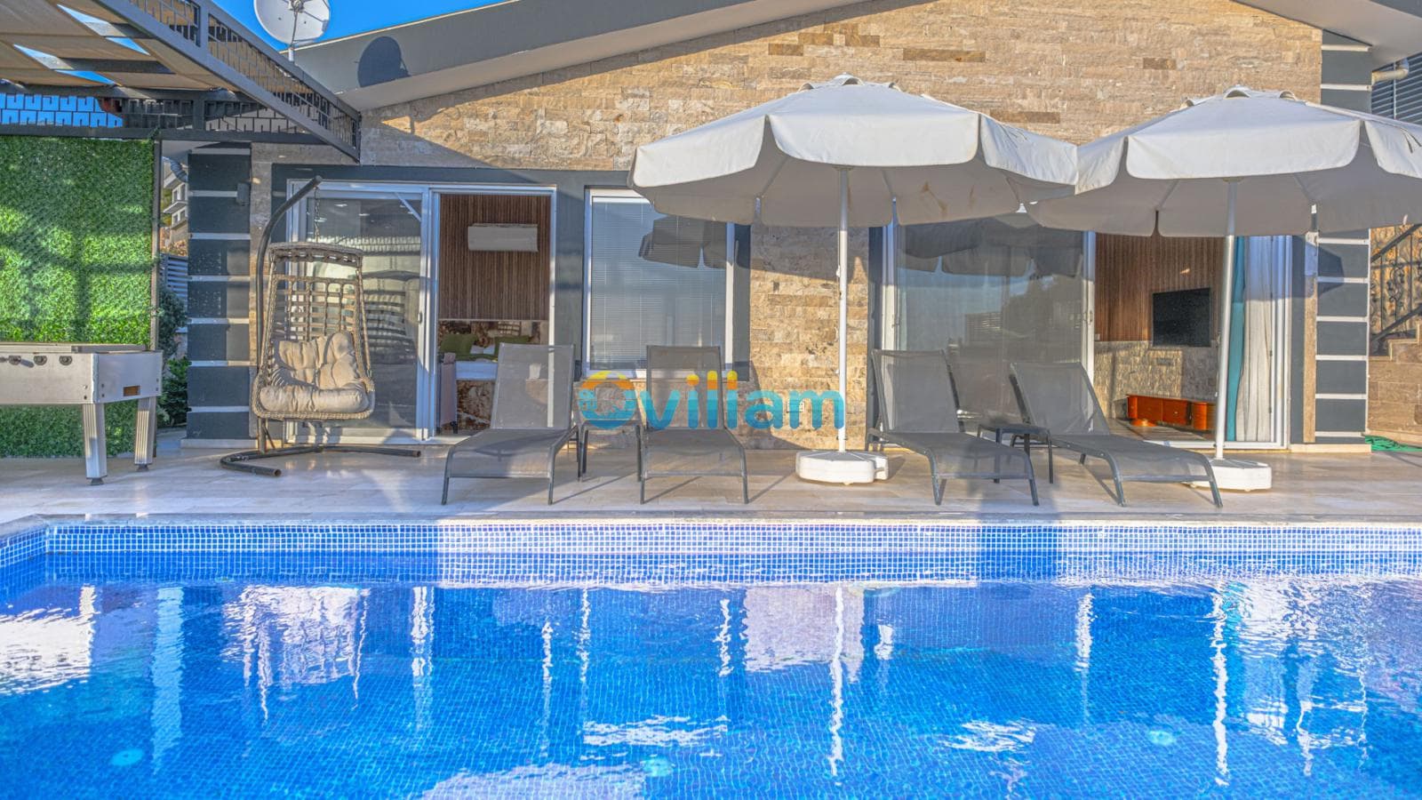 Havuz kenarında şezlonglar, güneş şemsiyeleri, asılı salıncak ve taş duvarlı villa dış cephesi.