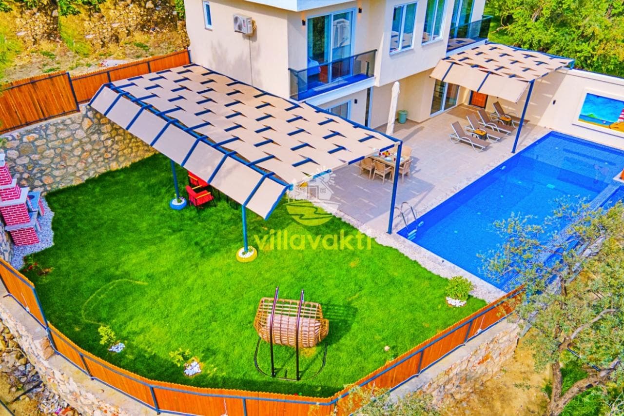 Havuz başında şezlonglar, yemek masası, gölgelikli oturma alanı ve çim bahçesi olan iki katlı villa.
