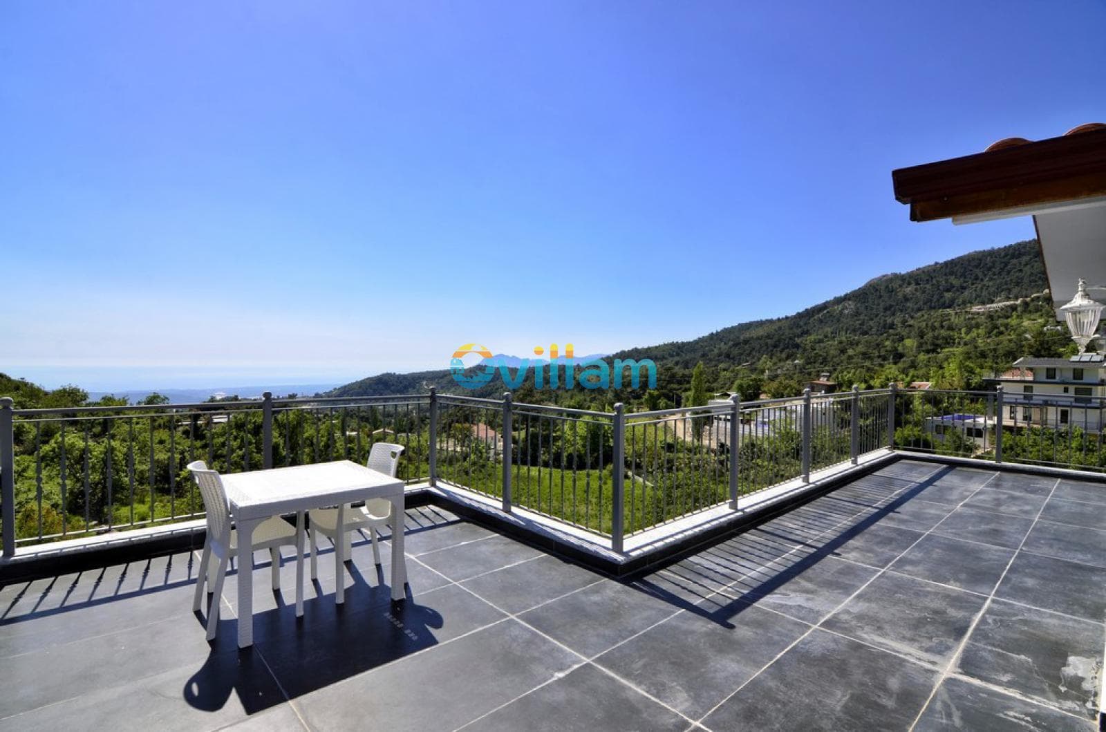 Dağ ve deniz manzarasına sahip, beyaz masa ve sandalyelerin olduğu geniş bir balkon.