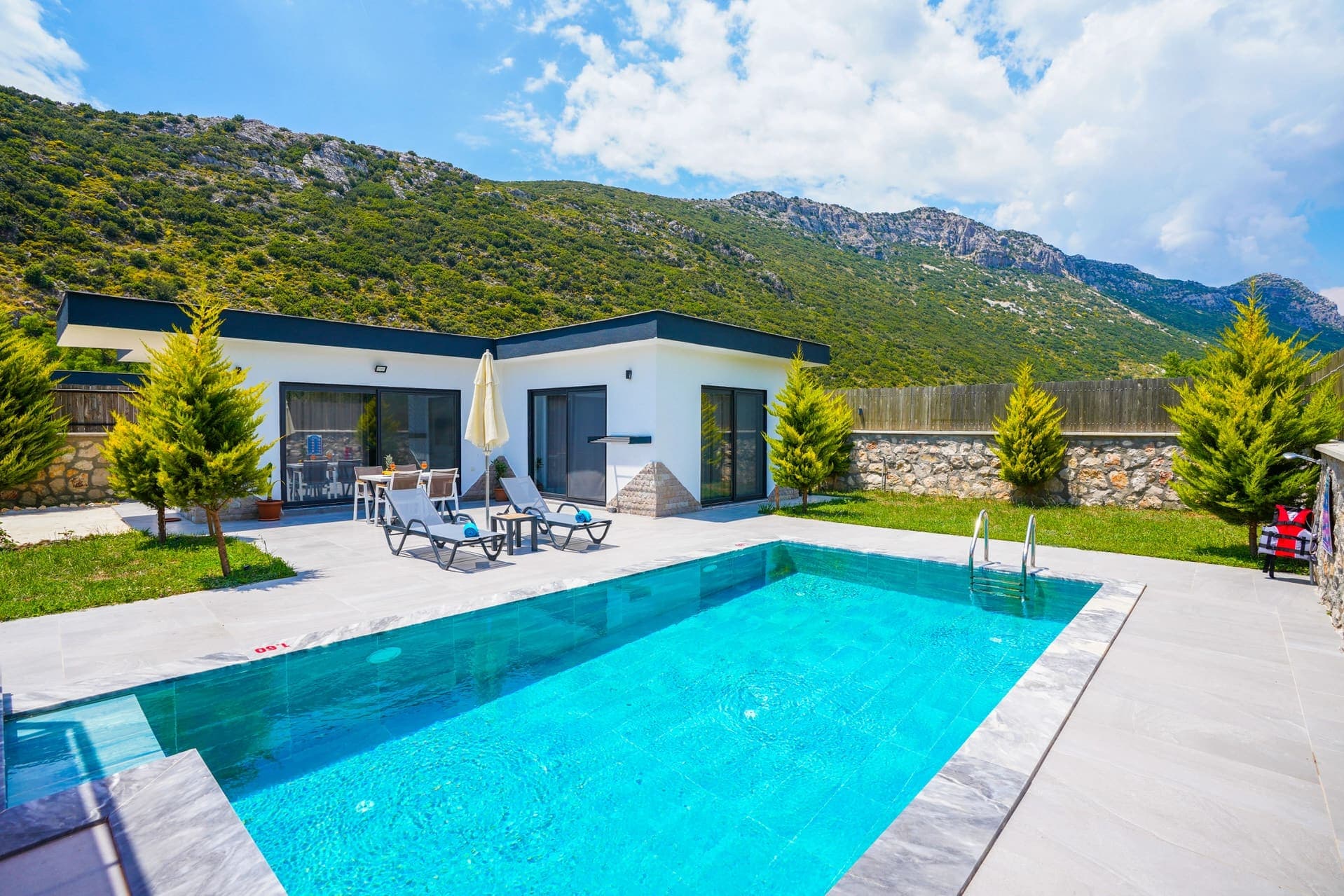 Dağ manzaralı, bahçesinde özel yüzme havuzu ve şezlonglar bulunan beyaz dış cepheli villa.
